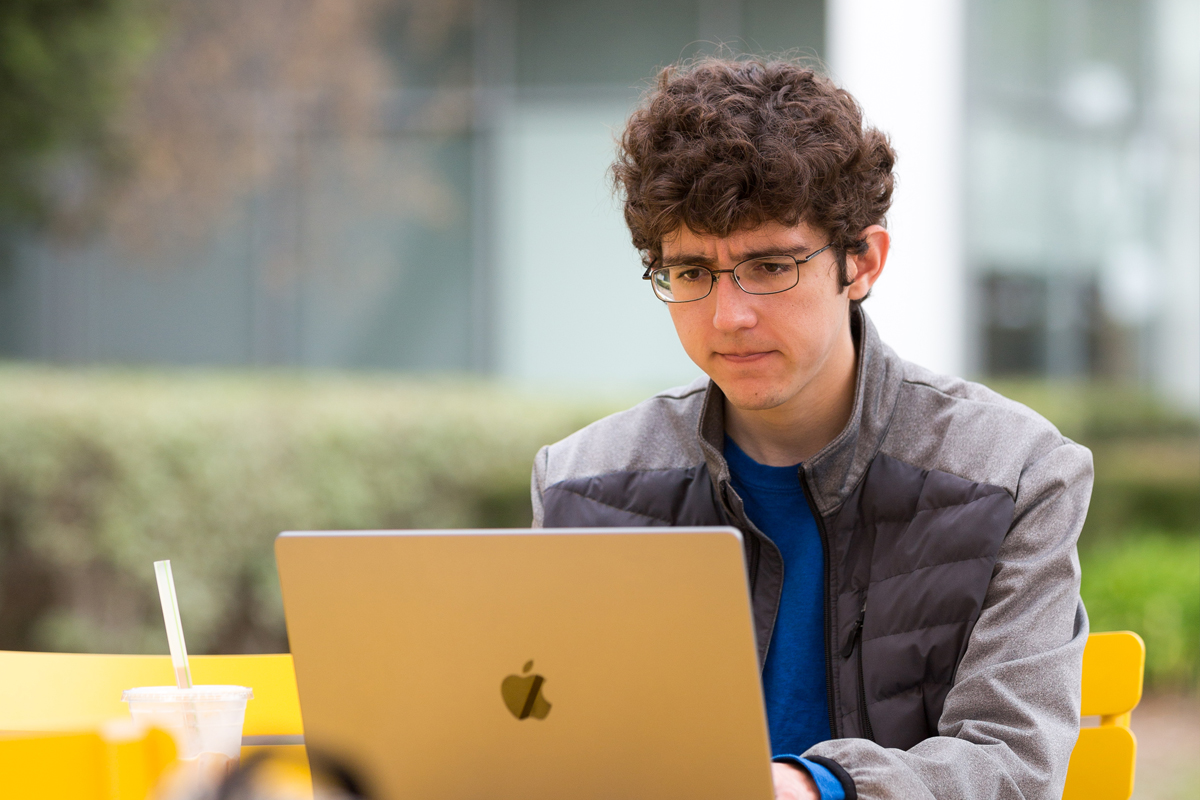 A student working on a laptop, outside at a picnic table.